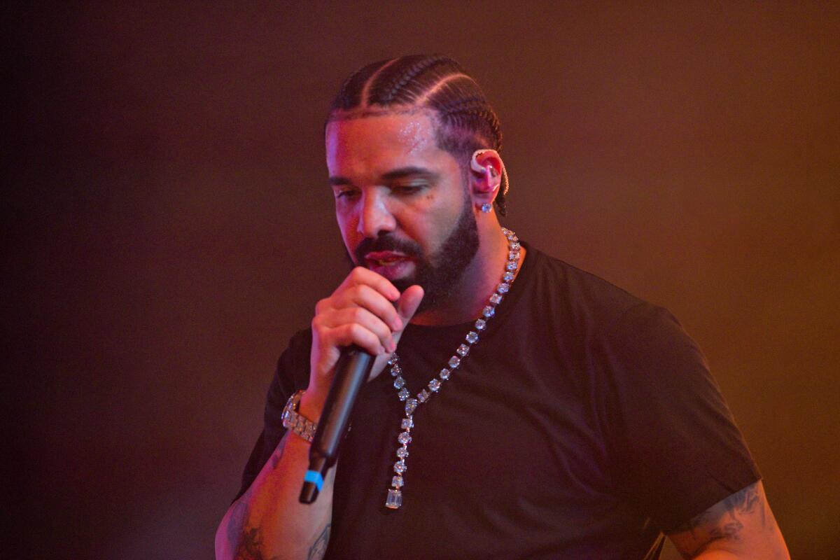 Drake is performing on stage with eyes down and a mic to his mouth while wearing a black T-shirt and diamond necklace