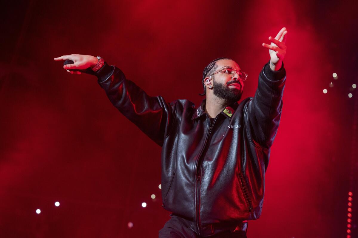 Drake in a large black leather jacket and glasses holding his hands out in front of him on a stage