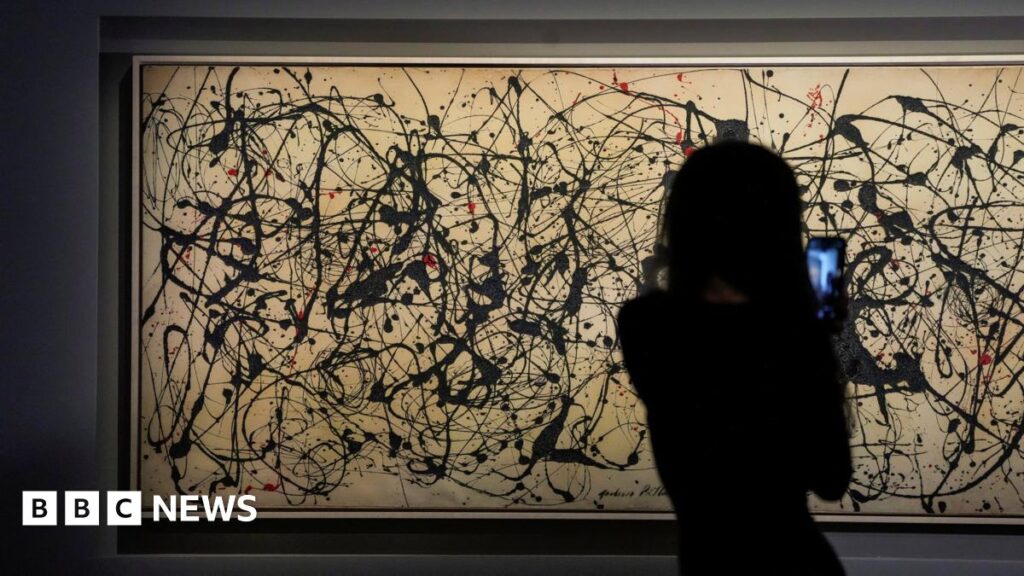 Jackson Pollock painting sells for record $181m at US auction A person with long hair in silhouette is seen facing the artwork holding their mobile phone to video the painting. The artwork is long horizontally and the grey canvass appears a light tan colour under the gallery lights. It features black paint flicks and splashes all over it, together with some fewer red markings.