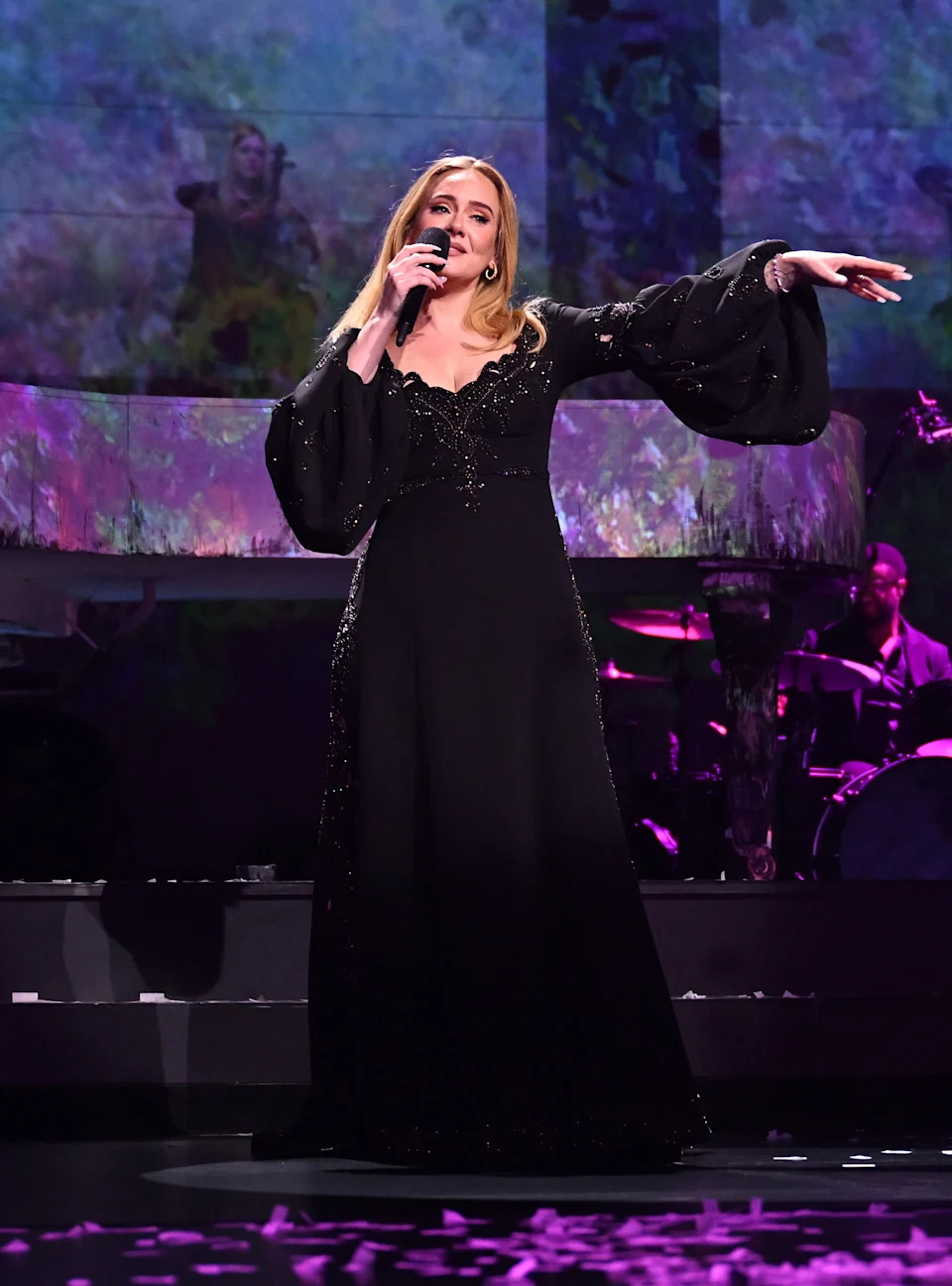 Singer performing on stage in a flowing black gown, holding a microphone, with musicians and a backdrop of nature imagery behind