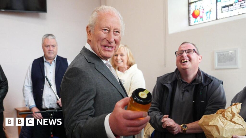 The King wears a dark-grey suit, white shirt and grey tie. He holds a clear jar of honey in his left hand. Her laughs as three people stand behind him. They are inside a building with white walls. A TV is over the King's right shoulder and a stain-glass window is over his left.