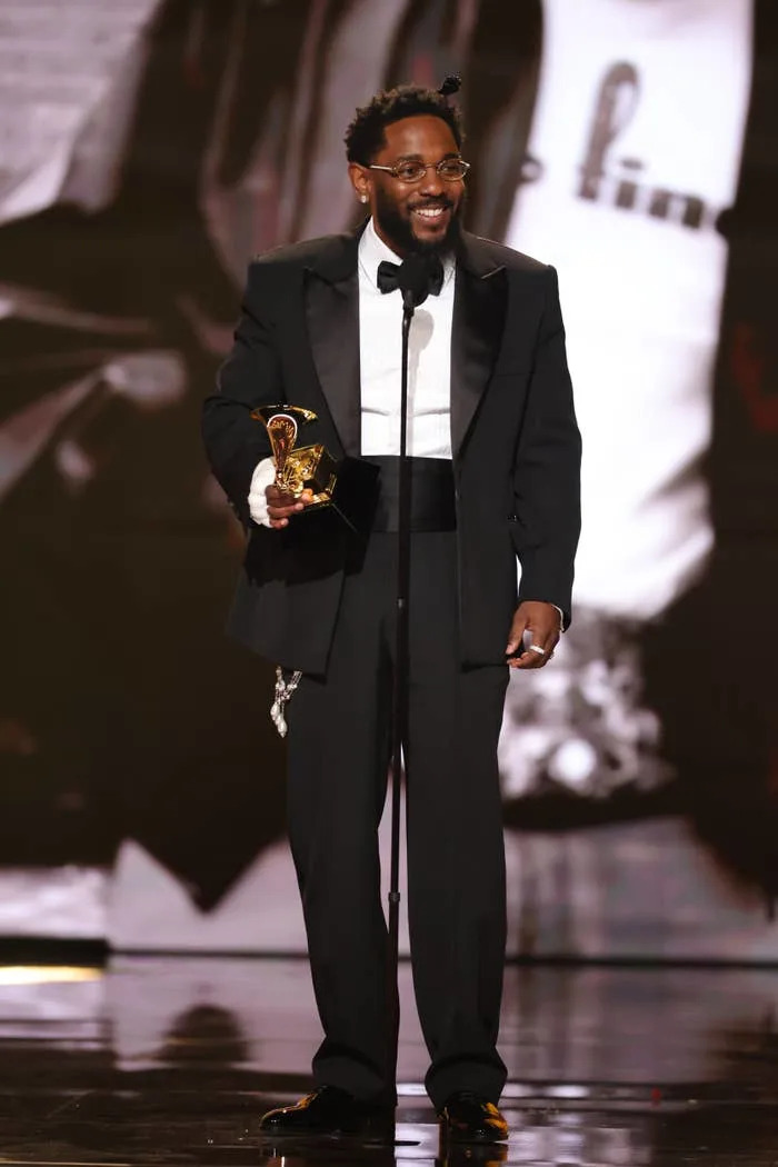 Person in a formal suit on stage holding an award, smiling at a microphone stand