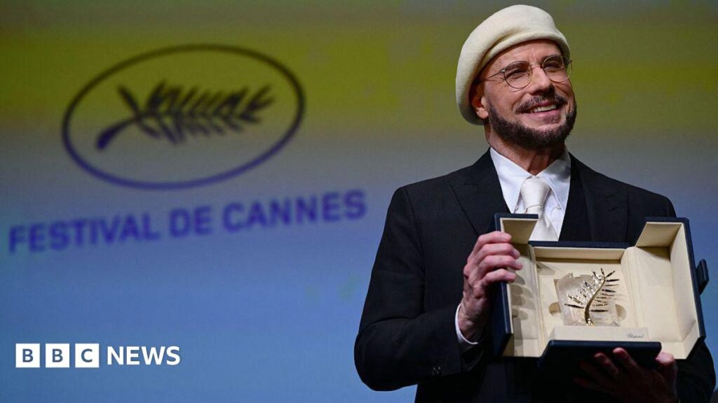John Travolta holds a sparkling award in box while smiling. He is wearing circular glasses and a white baret with a black suit. In the background is a green and blue screen with the words "Festival De Cannes" written on it.