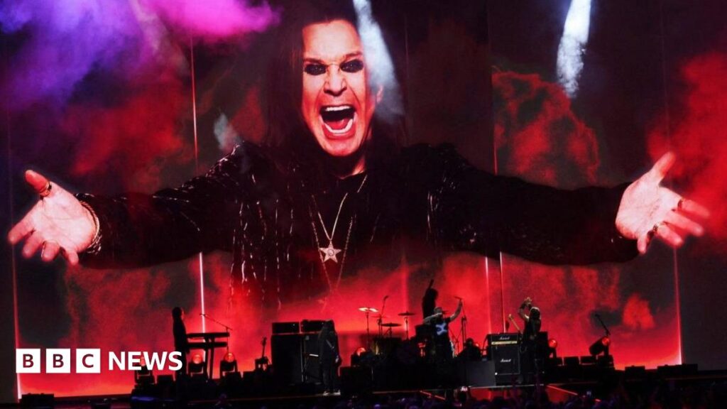 A band can be seen in red and black silouhette at the bottom of the image. On top the image of a man wearing black, chains around his neck, arms outstretched, dark eye make up and with a big smile