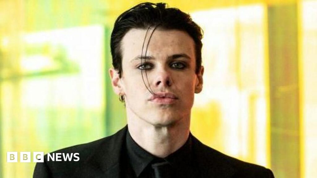 Yungblud stalking suspect from Sheffield fails to turn up at court Yungblud looks straight at the camera, wearing a black suit and black tie, against a blurred background.