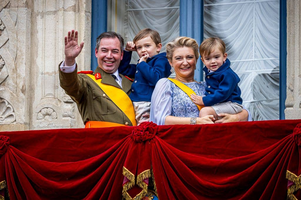 Grand Duke Guillaume waving while standing with Grand Duchess Stephanie on a balcony. The pair are holding their young children