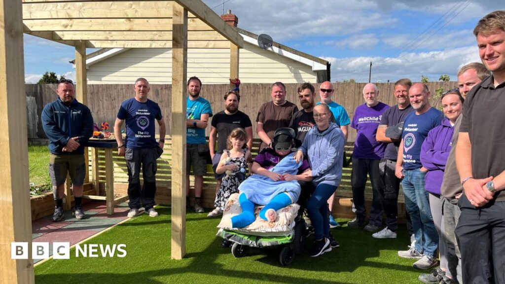 A group of people standing in an astroturf area smiling and looking at the camera. They are surrounding a family of 4, 2 adults and 2  children, one of which is Lucas who is in his wheelchair in the garden.