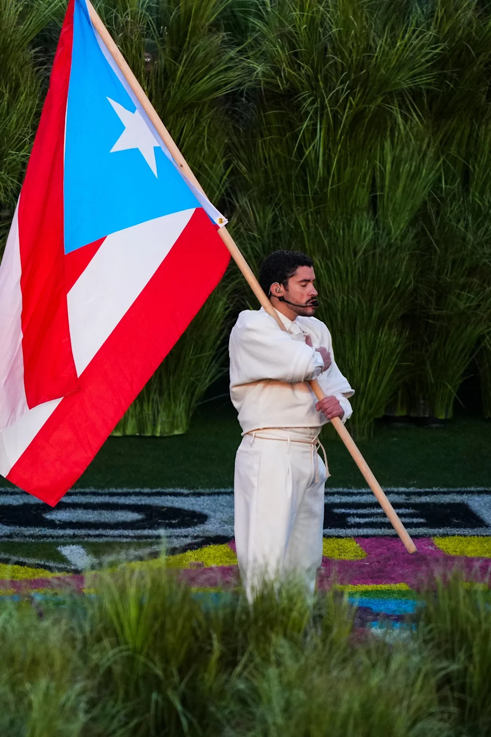 Person in cream suit holding Puerto Rican flag, standing on grass with greenery backdrop