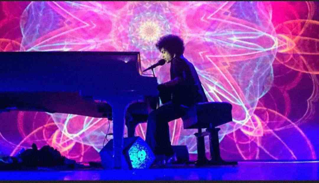 A musician with curly hair plays a grand piano and sings into a microphone, with vibrant, colorful, kaleidoscopic patterns projected in the background.