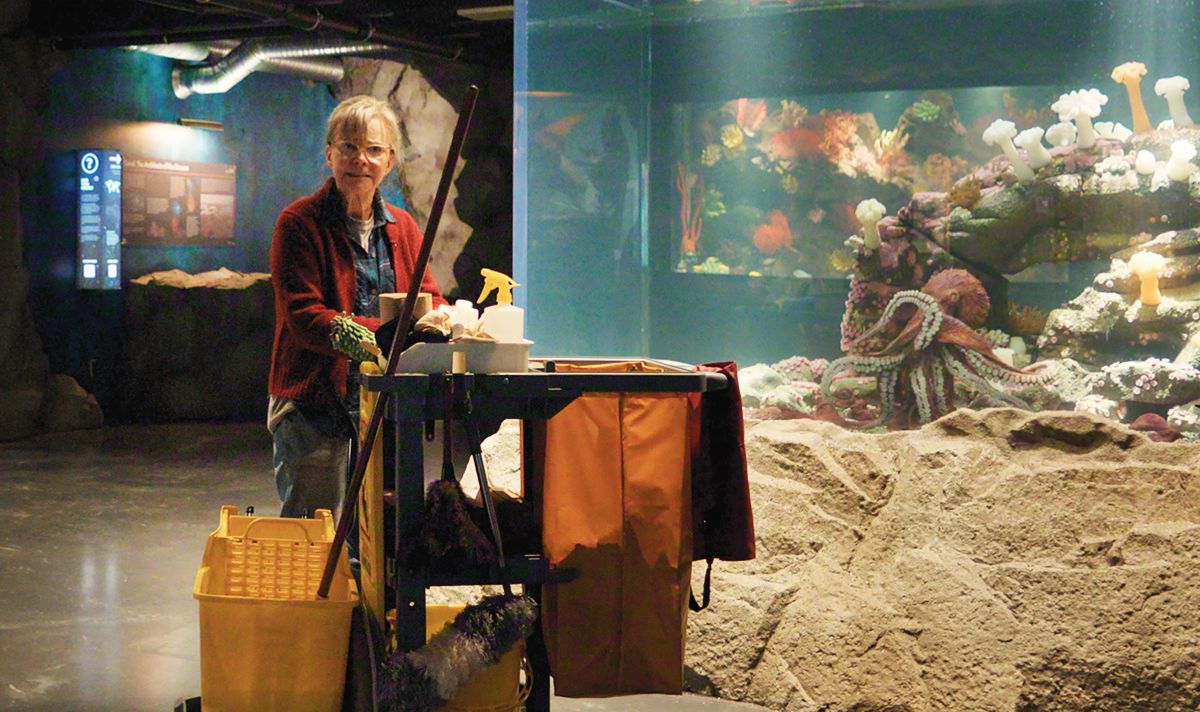Older woman in red cardigan with a cleaner's trolley by a large aquarium fish tank housing a red octopus