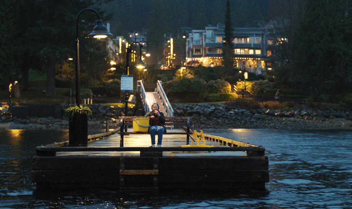 Woman sits on a pier next to a large yellow bucket at night with a lit-up town behind her