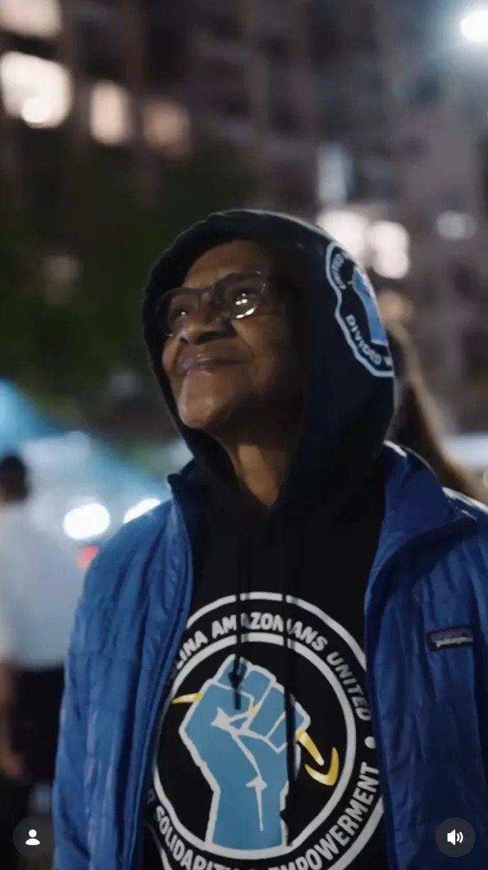 Person wearing a hooded jacket with a logo of a raised fist, looking up and smiling in an urban setting