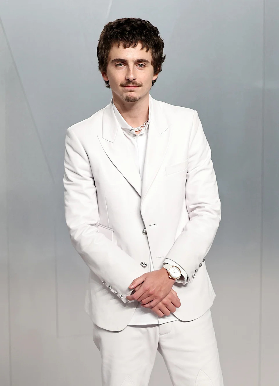 Person in a white suit posing with hands together, wearing a necklace and watch, against a plain background
