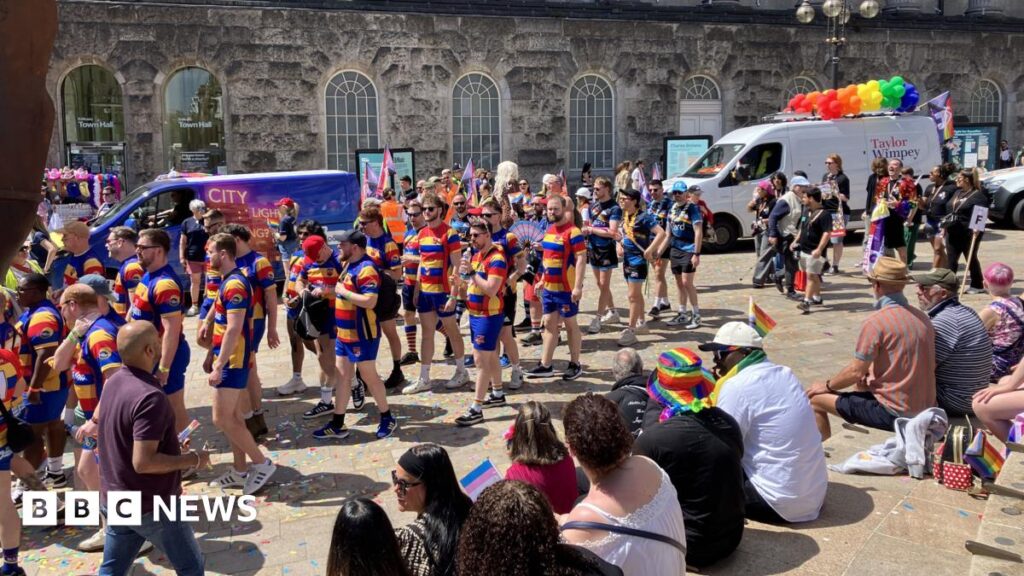dozens of people taking part in a parade. the people in the picture are dresses in blue short with blue, red and yellow striped tops. There is a van in the background with rainbow balloons strapped to its roof. People are sitting on nearby steps with rainbow flags
