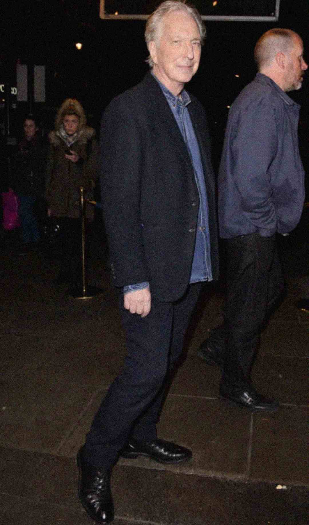A man with gray hair, dressed in a dark blazer, blue shirt, and black pants, stands and smiles slightly on a city sidewalk at night. Another man and several people are in the background.