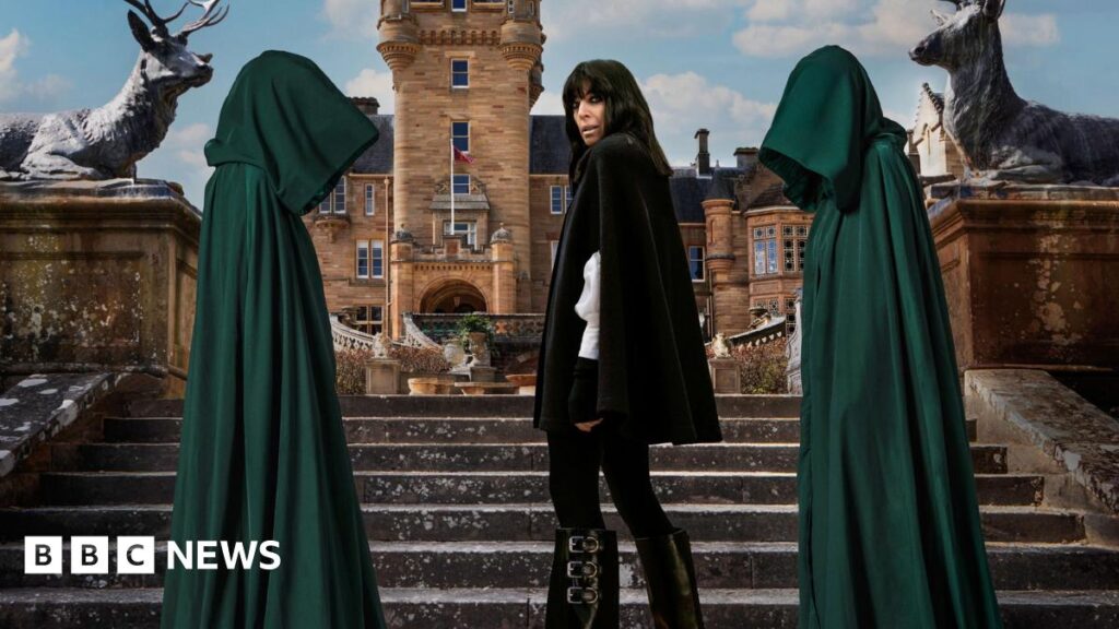 Claudia Winkleman stands on the castle steps, flanked by two hooded figures in green cloaks.