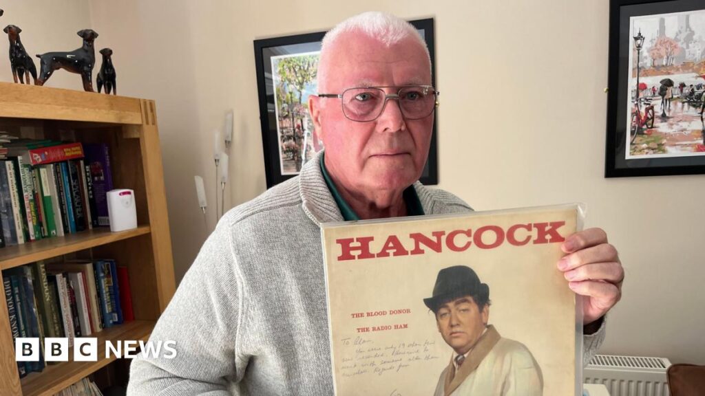 Alan Florence, has very short cropped grey hair he has silver framed glasses and is wearing a beige woollen cardigan with a green shirt beneath with the top buttons undone, behind him is a bookcase with vinyl albums and books and a small telescope in front of it.