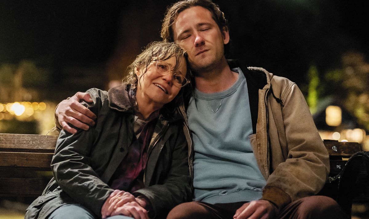 Older smiling woman and young man with his eyes closed sit on a bench, the man's arm around the woman