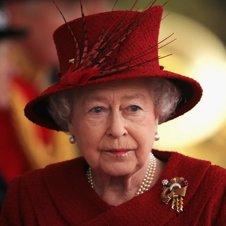  Queen Elizabeth wears a scarlet coat and matching hat with feathers in it and looks tearful. 
