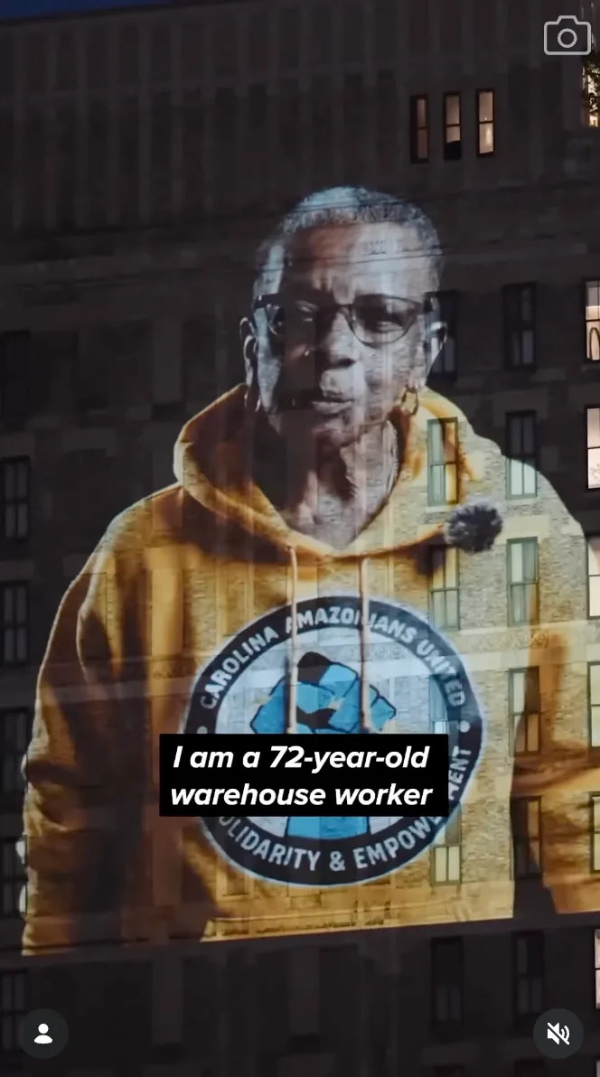 Projection of a person wearing a hoodie with a "Carolina Amazonians United" logo on a building, text reads: "I am a 72-year-old warehouse worker."