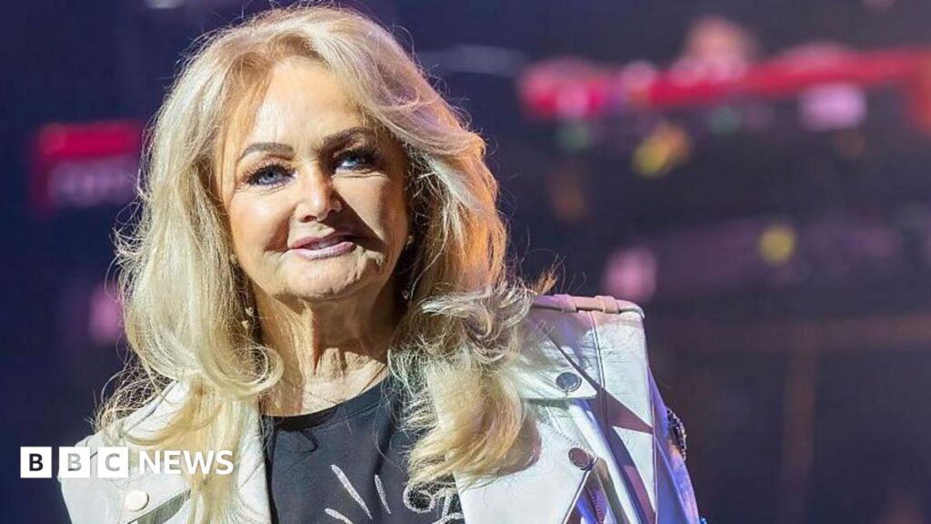 Bonnie Tyler ‘stable’ in Portuguese hospital Bonnie Tyler on stage, she has long blonde hair, and is wearing a black T-shirt and a white biker jacket