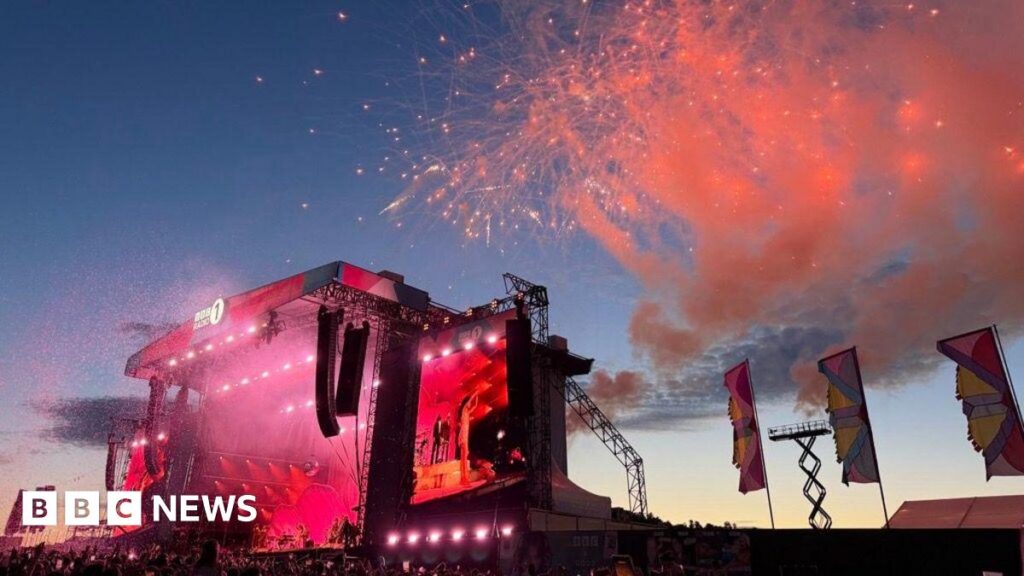 Olivia Dean is seen on stage on a screen, with pink lights and smoke in the sunset.