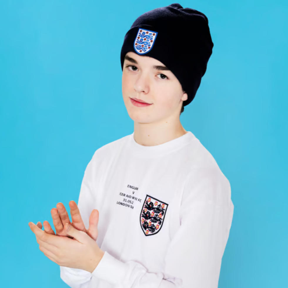 Soccer aid line up person with a beanie and longsleeve shirt clapping against a blue background