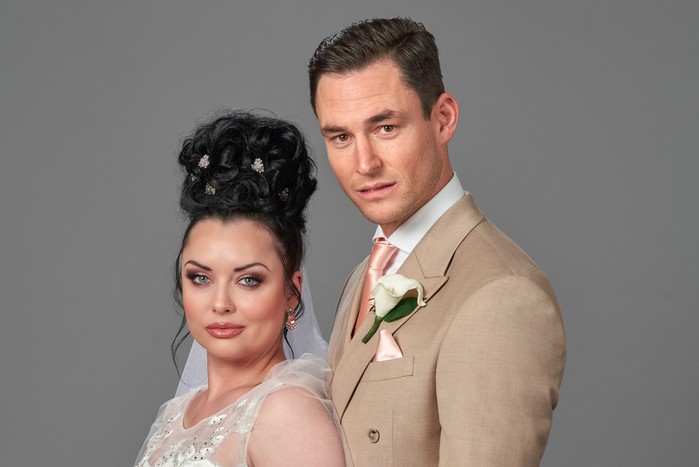 Whitney and Zack are posed facing the left, but looking into camera. Whitney (left) is in a wedding dress. Her hair is tied up decoratively above her head. Zack (right) is wearing a light beige suit. They're holding hands.