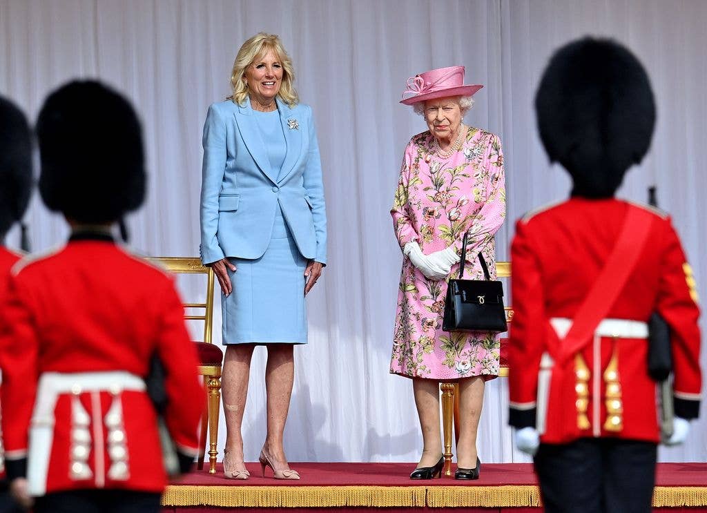 Jill Biden in blue suit and Elizabeth II in pink floral dress stand on stage with guards.