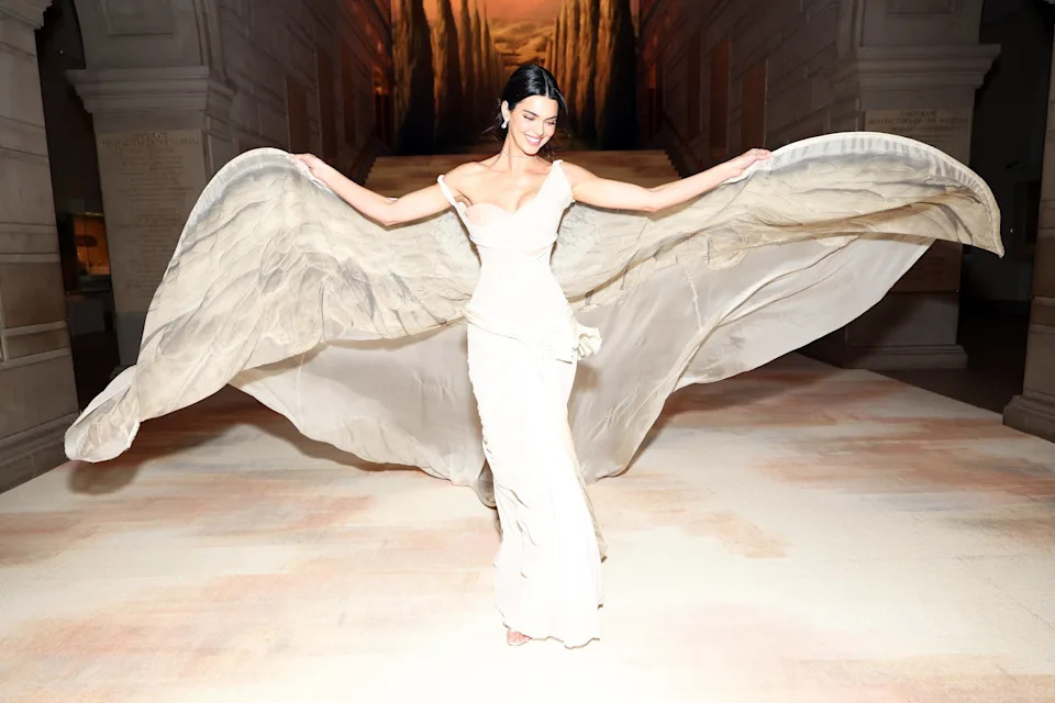 Person in elegant floor-length gown with flowing sheer train, posing indoors with arms outstretched, creating a dramatic wing-like effect