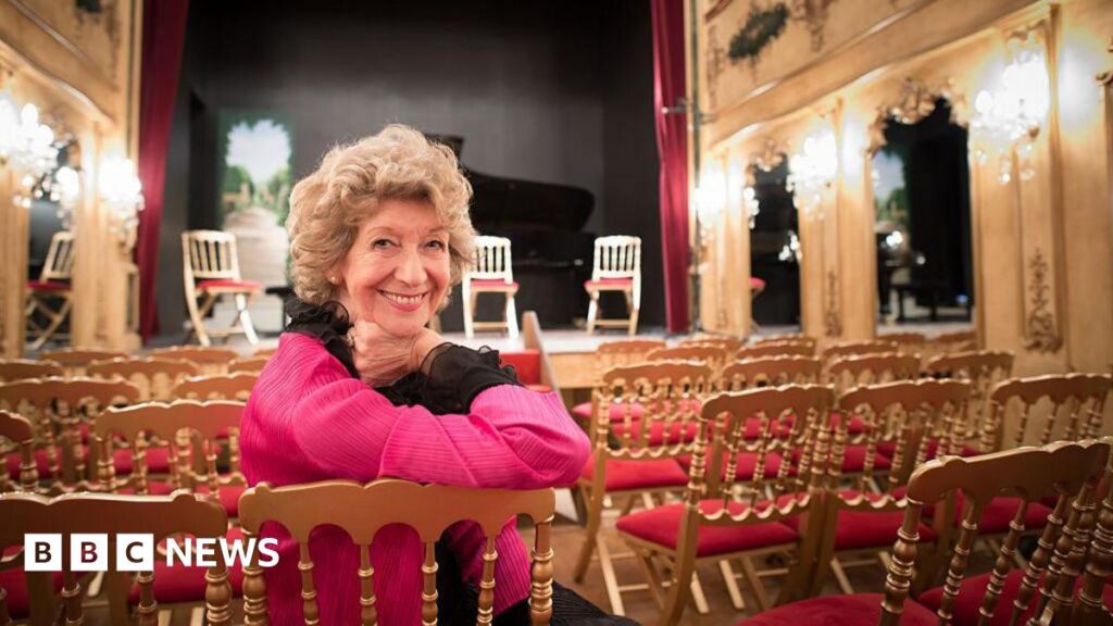 Dame Felicity Lott with short curly hair dressed in black and pink leans on the back of a chair and smiles to a camera while sitting in a theatre space.
