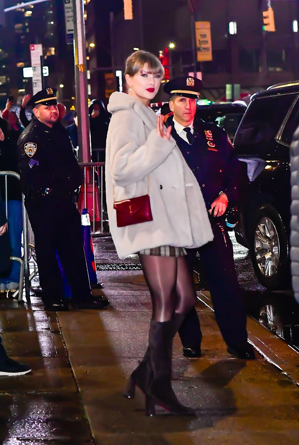 Person in a chic outfit with a short skirt, boots, and a plush coat waves while surrounded by police officers on a city street