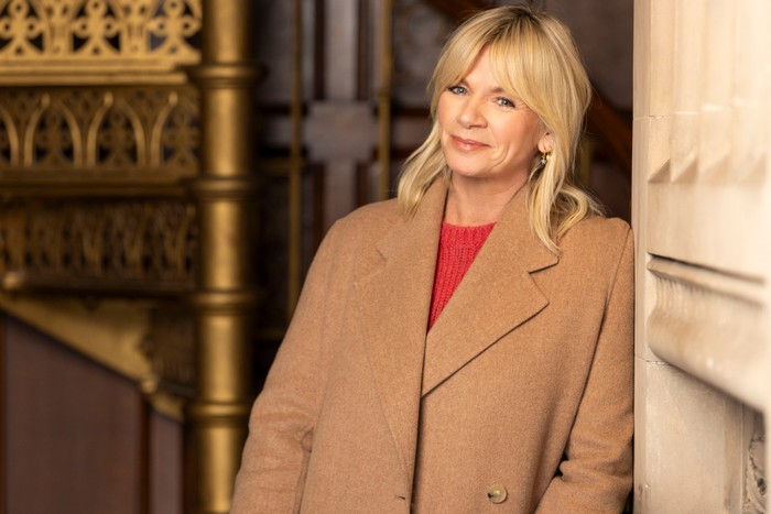 562217,TITLE:Who Do You Think You Are? s23 Zoe Ball stood resting her body to one side. wearing a red jumper and light brown jacket.