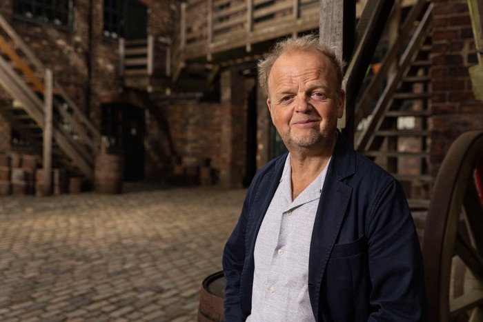 562218,TITLE:Who Do You Think You Are? s23 Toby Jones smiling ahead, wearing a white shirt and blue blazer.