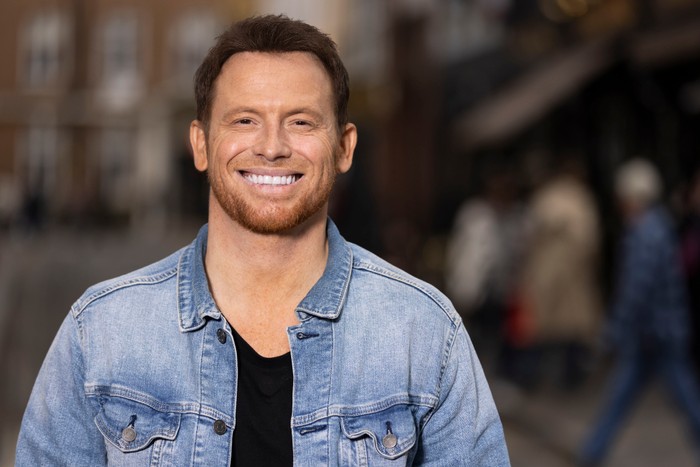 562220,TITLE:Who Do You Think You Are? s23 Joe Swash stood smiling ahead, wearing a black t-shirt and blue denim jacket.