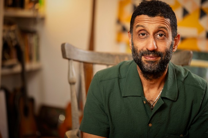 562224,TITLE:Who Do You Think You Are? s23 Adeel Akhtar sat on a chair, smiling ahead, as he wears a dark green shirt. He has black hair and a beard with grey colours.