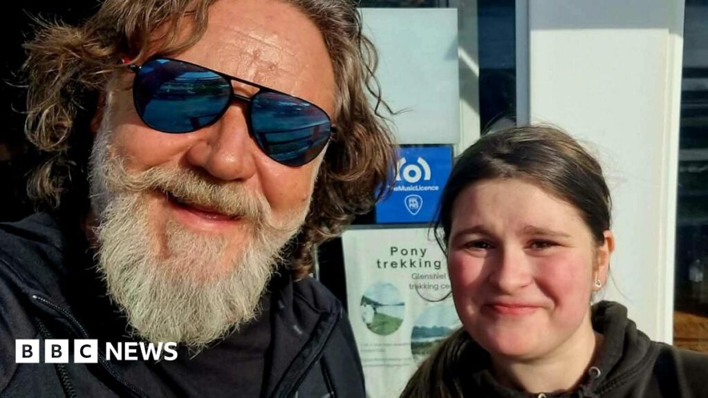 A man with a grey beard and shaggy brown hair. He's wearing mirrored sunglasses and is posing for a selfie with a young girl. She has dark hair and is wearing a black hoodie. They are both smiling.