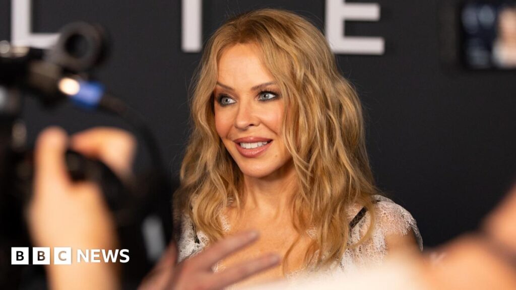 A woman (Kylie Minogue) with wavy blonde hair smiles while being photographed at an event, with blurred cameras and people in the foreground.