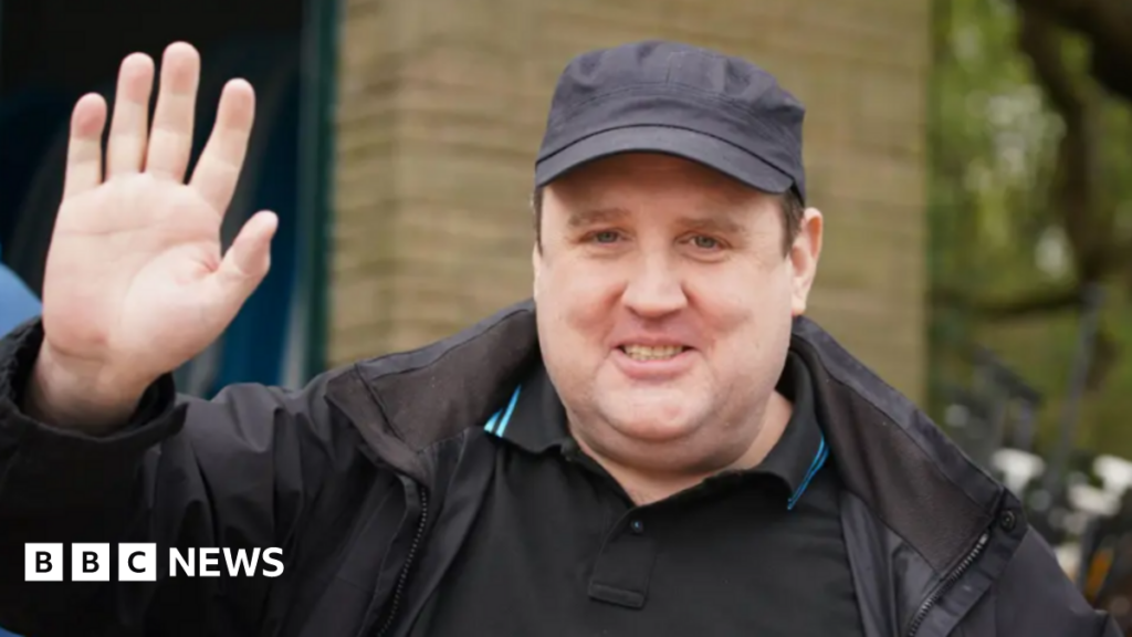 Kay is pictured waving at the camera and wearing a dark cap and top and jacket. He is outside in a street.