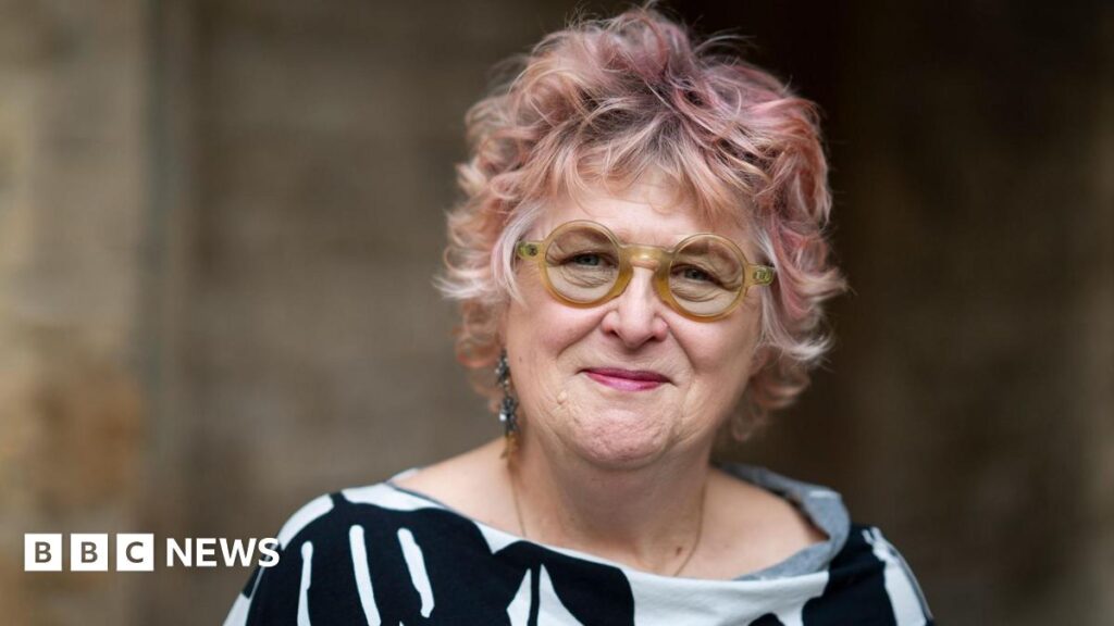 Close-up of a middle-aged woman with short pink hair and round green glasses. She is wearing a black and white checked scarf and looks sorrowfully at the camera.