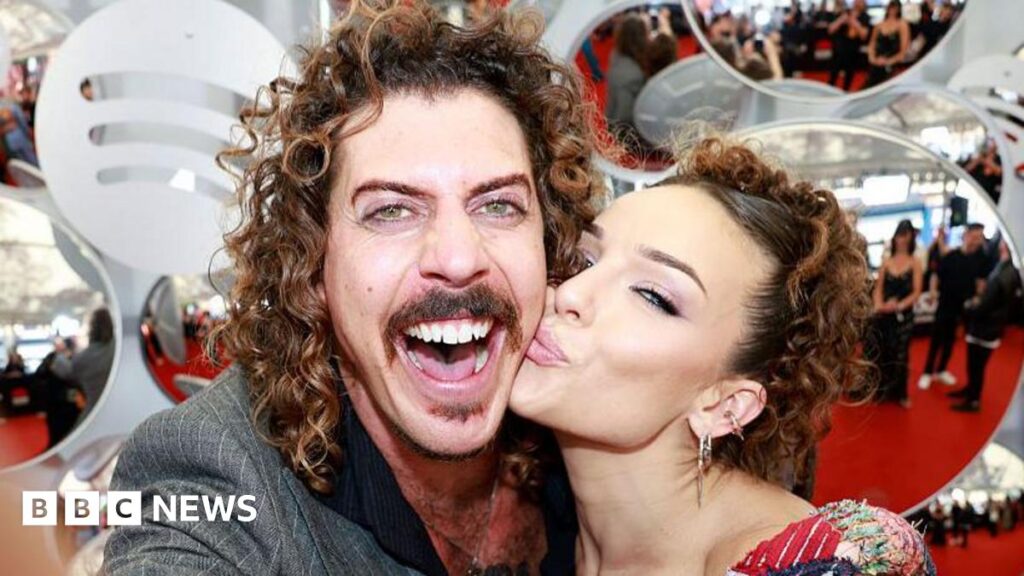 A man with curly brown hair, smiling widely at the camera, while a woman kisses him on the cheek
