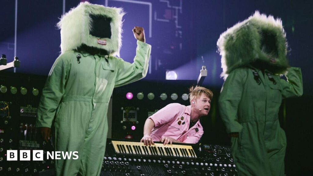 A man in a pink shirt playing a keyboard. On either side of him are two people, dressed in a green jumpsuit and a television prop over their heads.