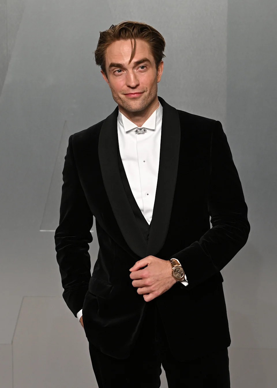 A man in a classic velvet tuxedo jacket and white shirt poses on a formal event backdrop