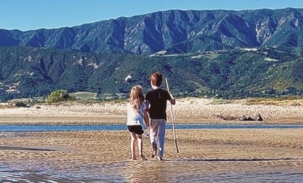 Two individuals, one holding a stick, are strolling on a sandy beach with a serene body of water in the background.