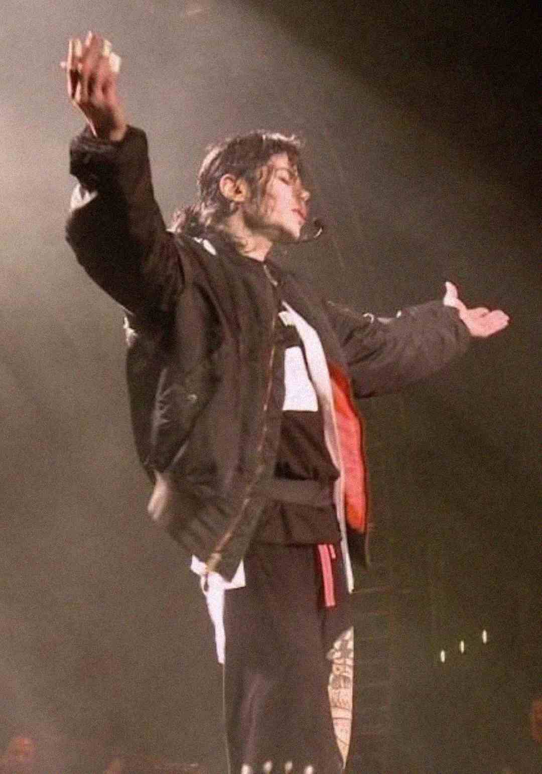 A person wearing a black jacket stands on stage with arms outstretched, facing to the side, under dramatic lighting.