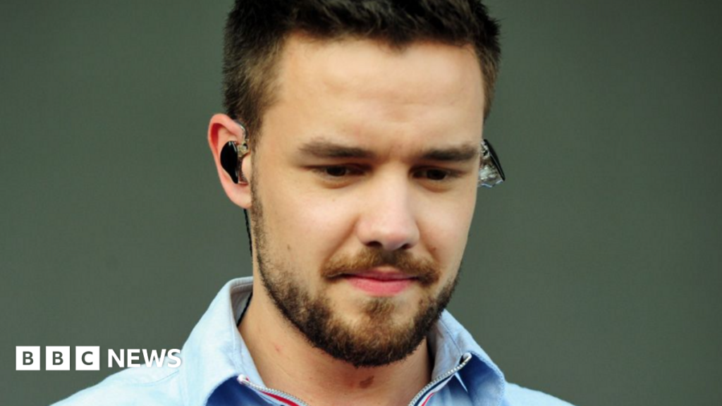 Liam Payne, with dark hair and a beard, is looking down and wearing an earpiece in front of a plain background.