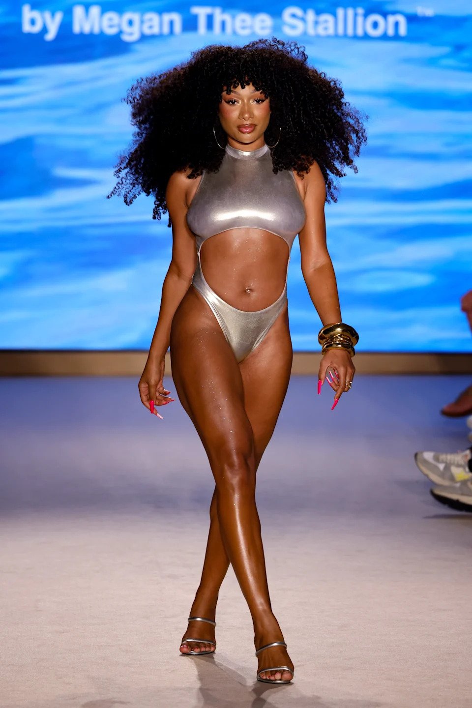 Model walks runway in metallic one-piece swimsuit with cutouts, hoop earrings, gold bangles, and heels. Blue backdrop with text partially visible