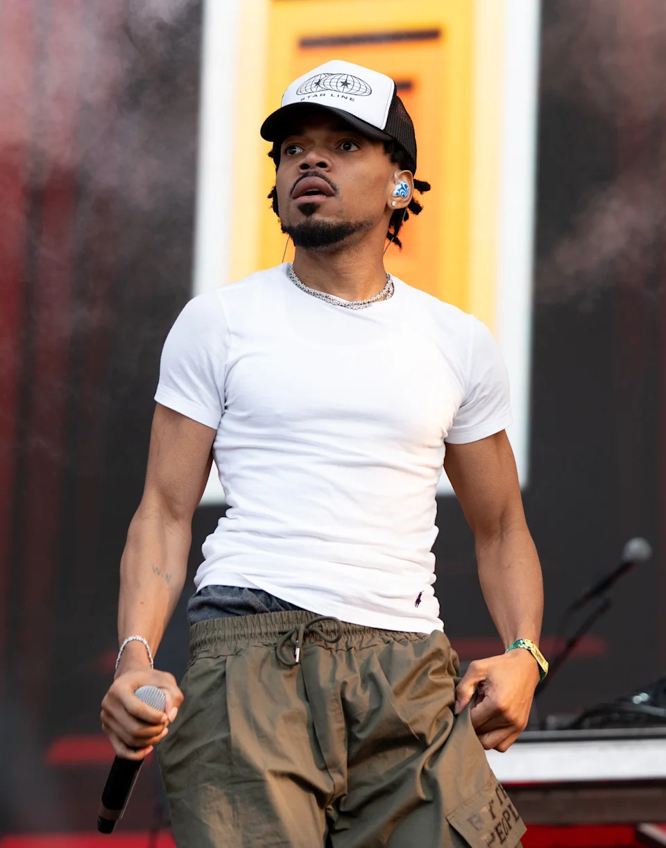 A person on stage holding a microphone, wearing a white t-shirt, cargo pants, and a baseball cap. They appear engaged in a performance