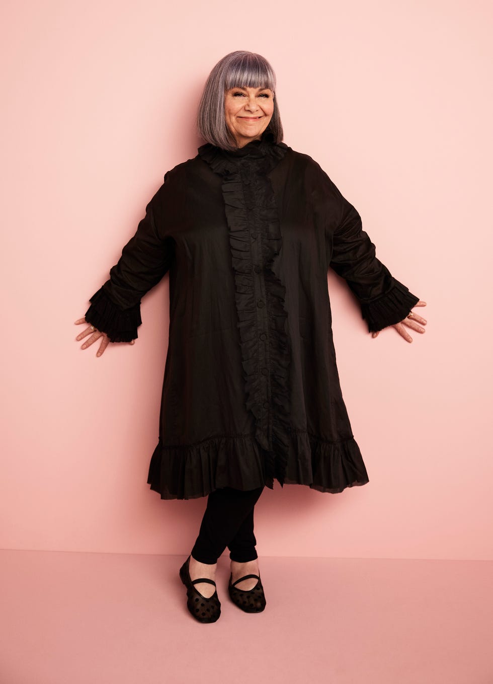 individual in a black ruffled coat with a light pink background