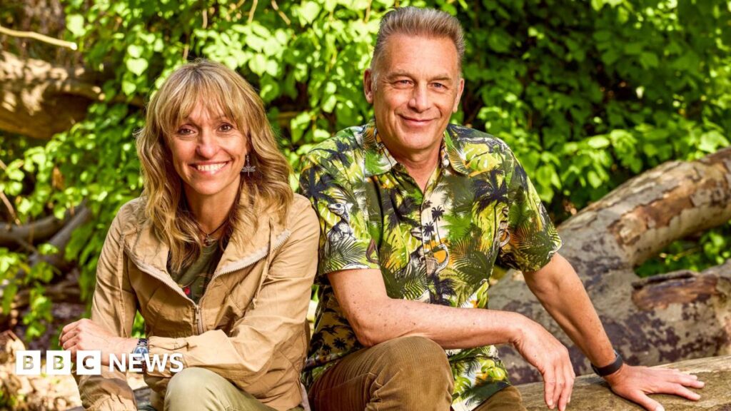 Michaela Strachan and Chris Packham are sitting outside on an old tree which is in a forest. They are smiling at the camera.